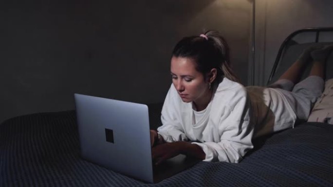 女人在现代酒店公寓吃早餐。窗灯肖像穿着浴袍的年轻女孩躺在床上用笔记本电脑工作。背景上有餐点、果汁、盘