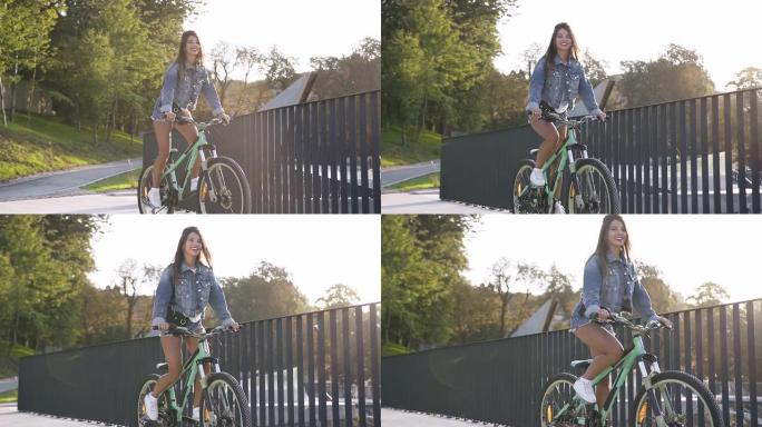 穿着牛仔裤衣服的可爱微笑辣妹骑在金属栅栏附近的自行车上