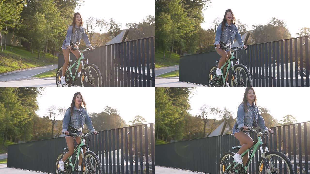 穿着牛仔裤衣服的可爱微笑辣妹骑在金属栅栏附近的自行车上