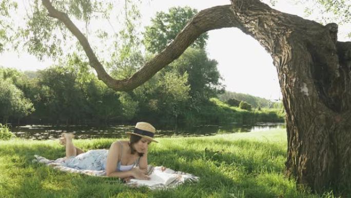 戴着交叉腿的帽子的女孩躺在河边大树下的草地上的格子上，读日记，在温暖的晴天放松和娱乐。在公园学习和阅