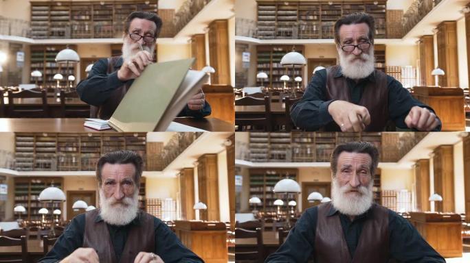 迷人的大胡子老人坐在图书馆的桌子旁，脱下眼镜，在相机上摆姿势合上书