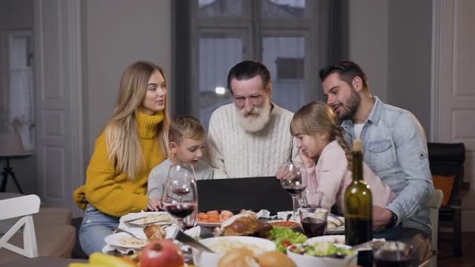 尊敬的祖父在餐桌上打开计算机，并与家人一起观看照片