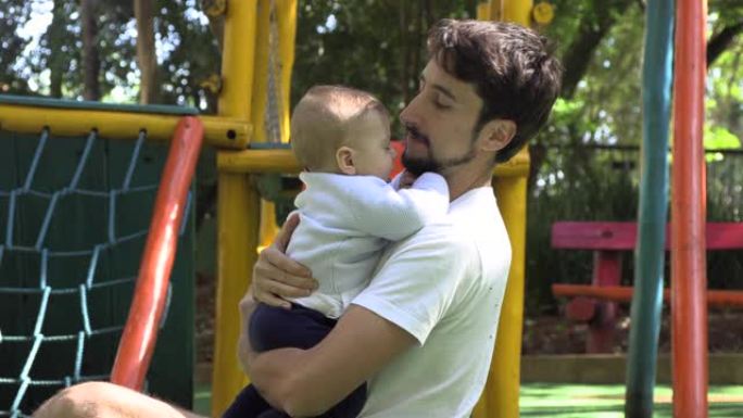 父亲在游乐场公园外面抱着婴儿儿子拥抱并充满爱心