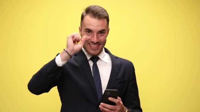 穿着西装的年轻时尚男子拿着手机阅读信息抬头微笑指着黄色背景的手指微笑