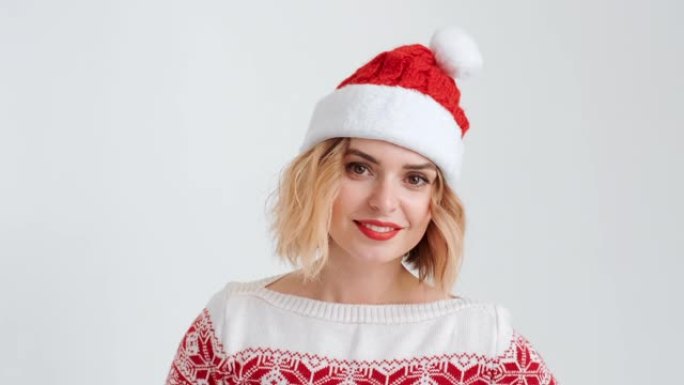 圣诞小红帽装扮的金发女郎面带微笑，随着音乐起舞，在白色背景上带着节日的气氛看着镜头。圣诞老人的帮手。