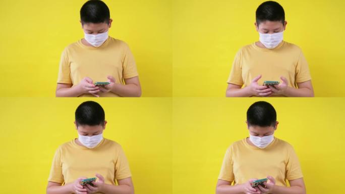 亚洲青春期男孩戴着面罩，使用黄色背景的智能手机，这是新的正常概念。