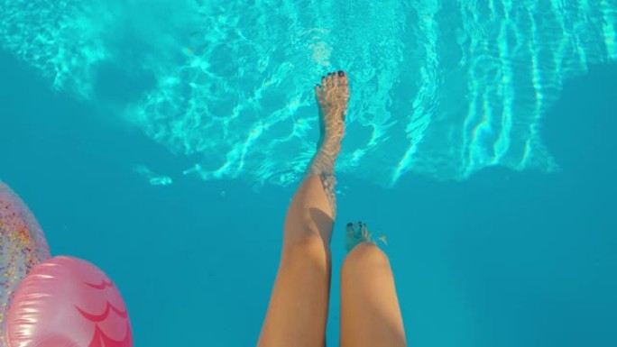 在炎热的夏日，在阳光下，苗条的女性双腿紧贴游泳池旁的蓝色水面特写。女孩在游泳池的水中晃来晃去，休息，