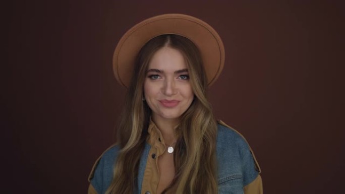 迷人的年轻女子在棕色背景上戴着棕色帽子对着镜头微笑