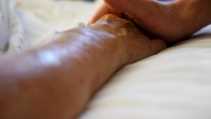 孙子在医疗诊所握住并安慰他老祖母的手。年轻人轻轻地抚摸躺在医院病床的生病成熟奶奶的皱纹手臂。儿子对母