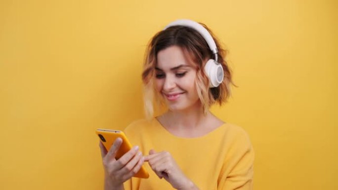 穿着白色耳机的快乐微笑的年轻性感女孩的肖像在智能手机中寻找好音乐，发现，在孤立的黄色背景上跳舞。乐事