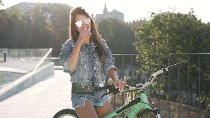 穿着牛仔裤的可爱快活性感女人诱人的衣服和时尚眼镜坐在自行车上，看着城市景观上的相机