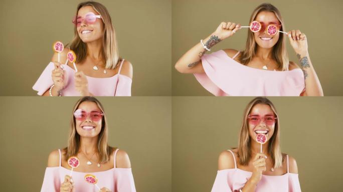 戴着太阳镜和粉色衬衫的年轻女士。她拿着两个葡萄柚棒棒糖，在橄榄工作室背景下摆姿势时微笑着跳舞