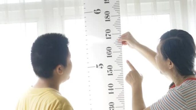 亚洲母亲对着窗户测量儿子的身高，生活方式的概念。