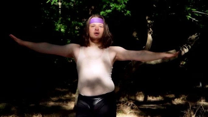 富有表现力的超重男人，长发，穿着有趣的女性衣服在森林里健身。顽皮的胖子描绘女孩做减肥体操。慢动作。
