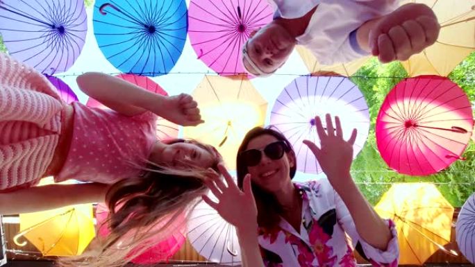 妈妈，爸爸和女儿三口之家在悬挂多色装饰伞的背景下自拍。他们在拍摄视频时跳舞并玩得开心