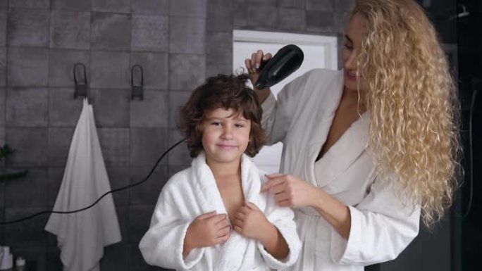 长发有爱心的年轻妈妈在浴室洗澡后，用吹风机吹干卷发她的小快乐儿子