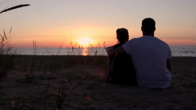 年轻的恋人夫妇在浪漫的金色日落时坐在沙滩上的沙滩上。后视图