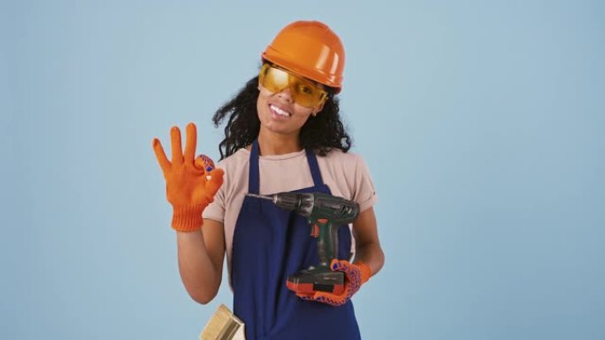 戴着安全帽、护目镜和手套的黑人黑人女孩工人。她拿着钻头，露出好的手势，微笑着。在蓝色背景上摆姿势