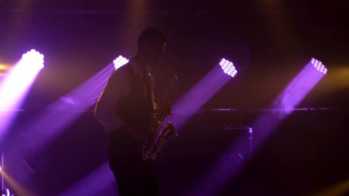 剪影一个年轻的时尚男人在舞台上的紫色聚光灯下演奏金色闪亮的萨克斯管。带烟雾和霓虹灯的黑暗工作室。动态