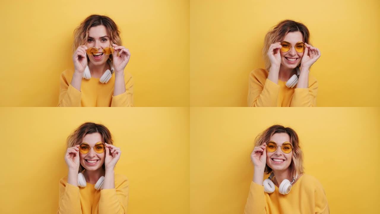 肖像快乐微笑的年轻性感女孩戴着白色耳机听音乐跳舞，在孤立的黄色背景上拉直黄色眼镜。乐事。人们的情感。