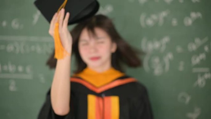 亚洲女毕业生穿着黑色长袍和黑色帽子，并配以金色流苏。站在教室的黑板前