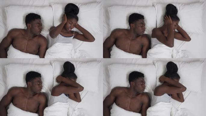 躺在床上的黑人夫妇的俯视图。不眠之夜的女孩用手遮住耳朵，转身回到她打呼噜的男朋友身边。
