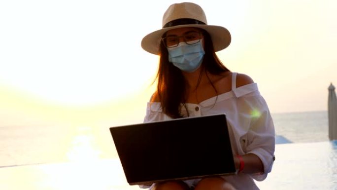 年轻女子戴着防护口罩，遮阳帽和夏装，在笔记本电脑上工作，坐在游泳池旁。远程工作。新常态。Covid1