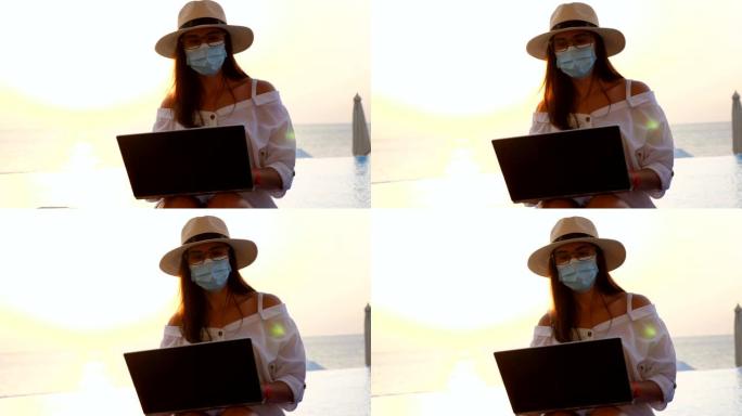年轻女子戴着防护口罩，遮阳帽和夏装，在笔记本电脑上工作，坐在游泳池旁。远程工作。新常态。Covid1