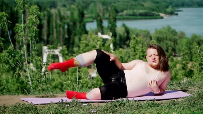 顽皮的胖子，长发，穿着紧身t恤和紧身裤，在户外做体操减肥。有趣的超重男孩描绘了一个在公园健身的女孩。