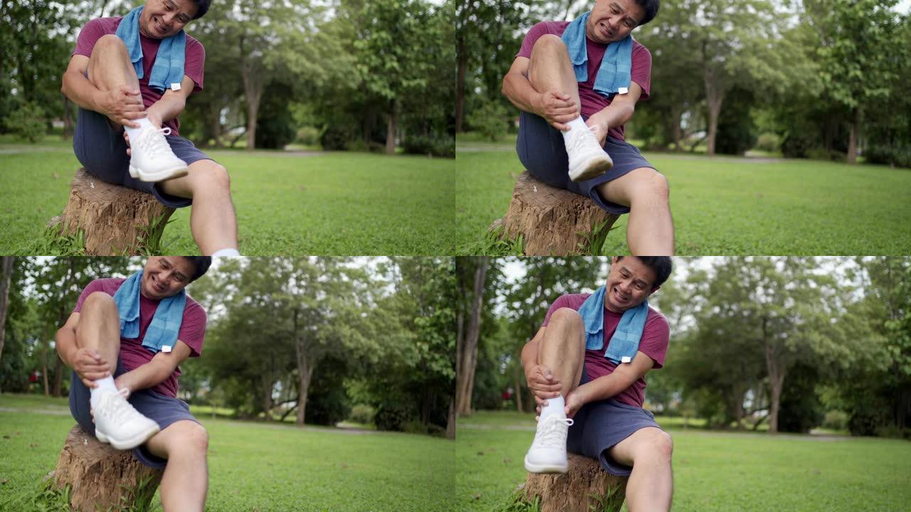 亚洲老年男性在公园内运动时扭伤脚踝坐在树干上，背景是草坪，脚踝疼痛，关节韧带问题，面部表情疼痛，事故