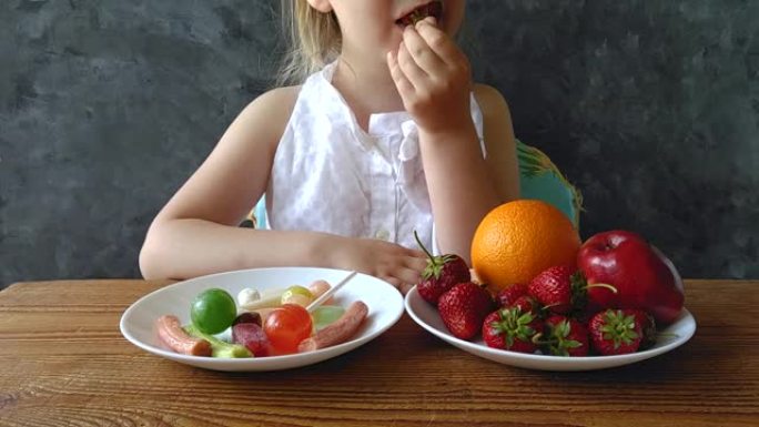 家里桌子上放着新鲜水果和糖果的小女孩。孩子认为选择健康或有害的食物。高清