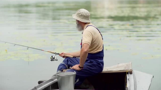 坐在木船上在湖里钓鱼的大胡子老渔夫的侧视中等镜头
