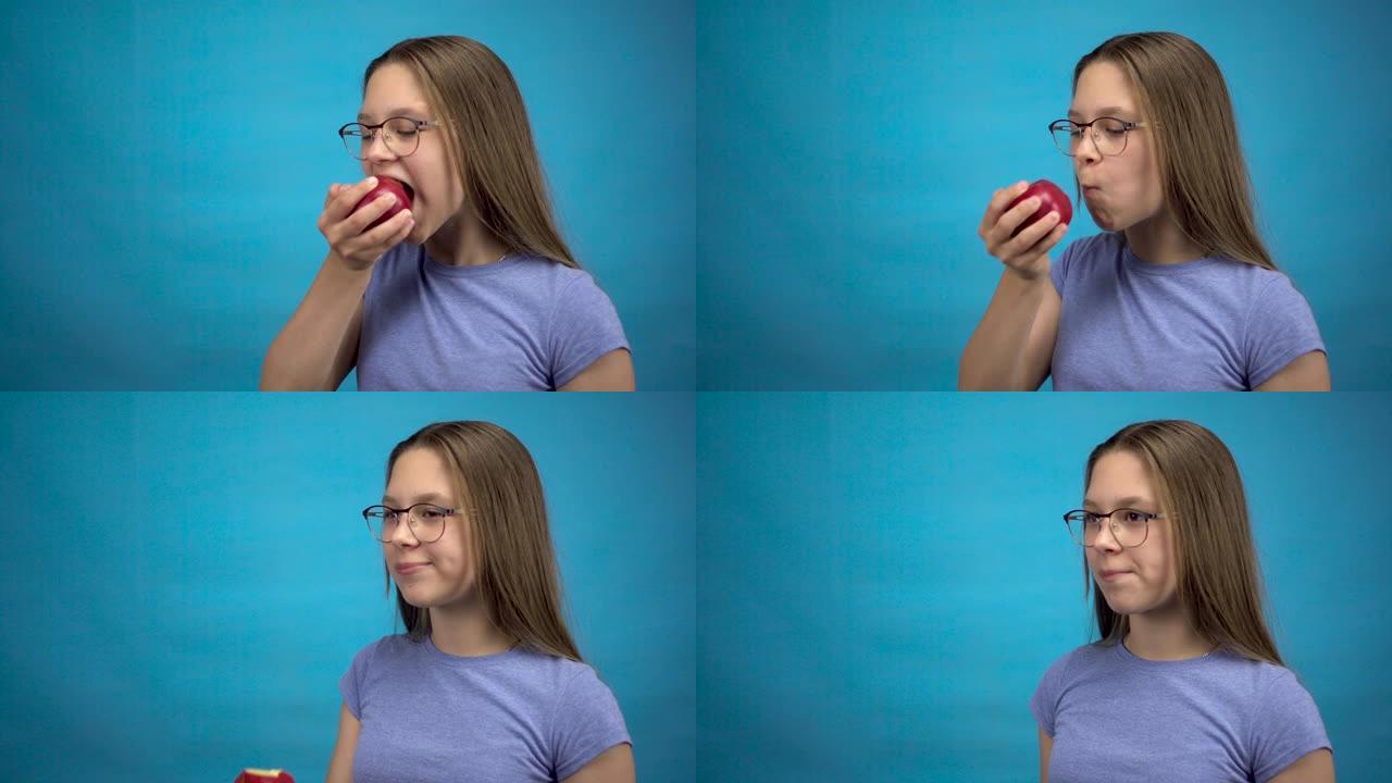 牙齿上有牙套的少女吃蓝色背景上的红苹果。戴着彩色牙套的女孩咬掉苹果。