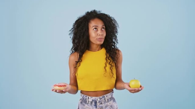 美国黑人模特从背后拿出苹果和甜甜圈，想着应该吃什么。选择甜甜圈，微笑。在蓝色背景上摆姿势