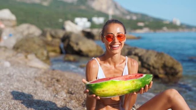 穿着比基尼的快乐女孩拿着大片西瓜坐在海边的沙滩上