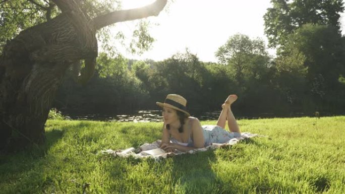 女孩在河边的草地上休息，享受温暖的晴天。穿着草帽的迷人黑发女人躺在河边大树下的草地上的格子上，看书日