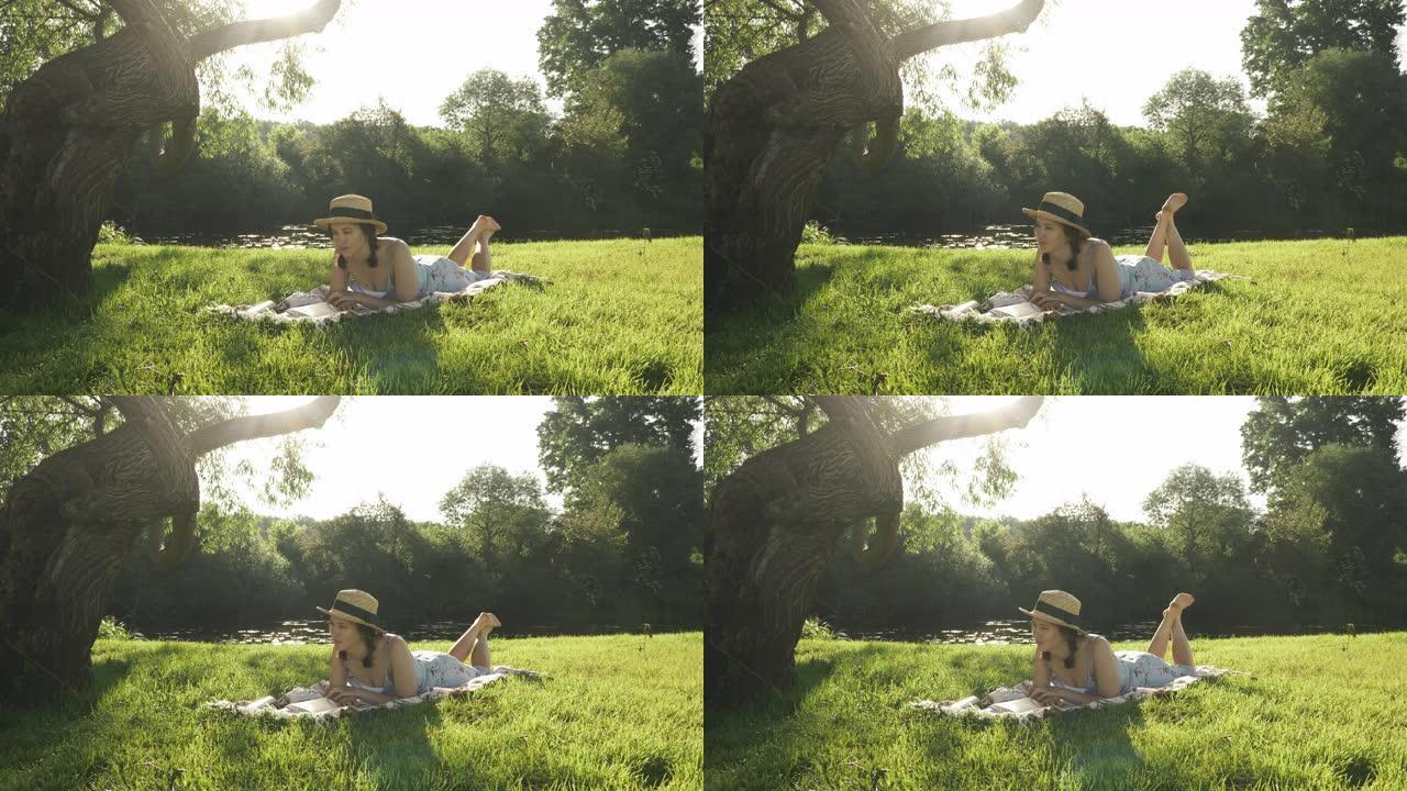 女孩在河边的草地上休息，享受温暖的晴天。穿着草帽的迷人黑发女人躺在河边大树下的草地上的格子上，看书日