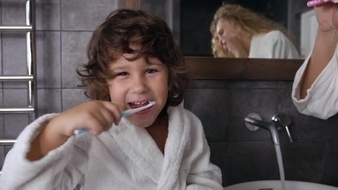 穿着白大褂的卷发开朗的小男孩正在浴室里用牙膏和牙刷一起给妈妈刷牙。多莉开枪