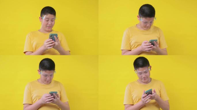 亚洲青春期男孩戴着面罩，使用黄色背景的智能手机，这是新的正常概念。
