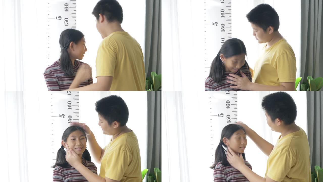 快乐的亚洲兄弟在家对着窗户测量他姐姐的身高，生活方式理念。
