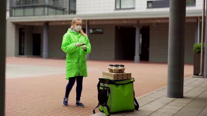 在防护面罩，手套和绿色制服中追踪女性食品快递员的照片将披萨盒和外卖咖啡杯放在保温袋上，并致电客户进行