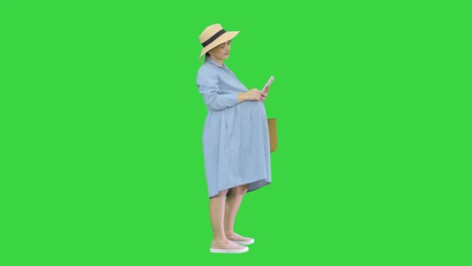 穿着蓝色连衣裙和帽子的快乐孕妇在绿色屏幕上使用智能手机，色度键