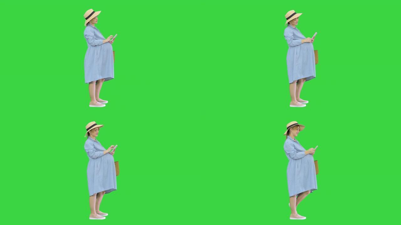 穿着蓝色连衣裙和帽子的快乐孕妇在绿色屏幕上使用智能手机，色度键