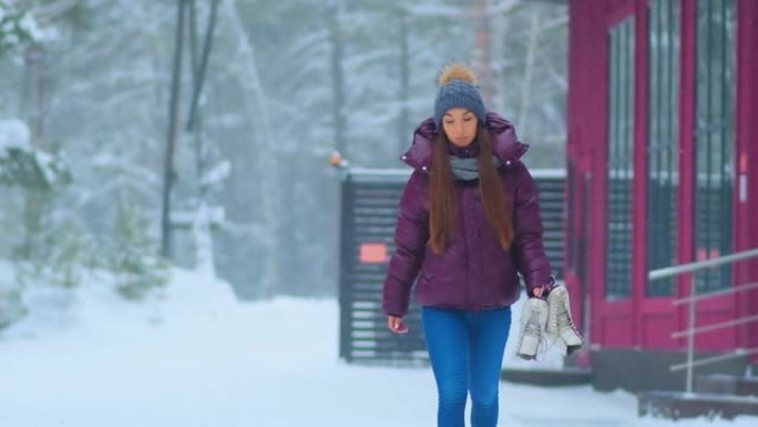 穿着紫色夹克的女人在白雪皑皑的公园里拿着溜冰鞋散步