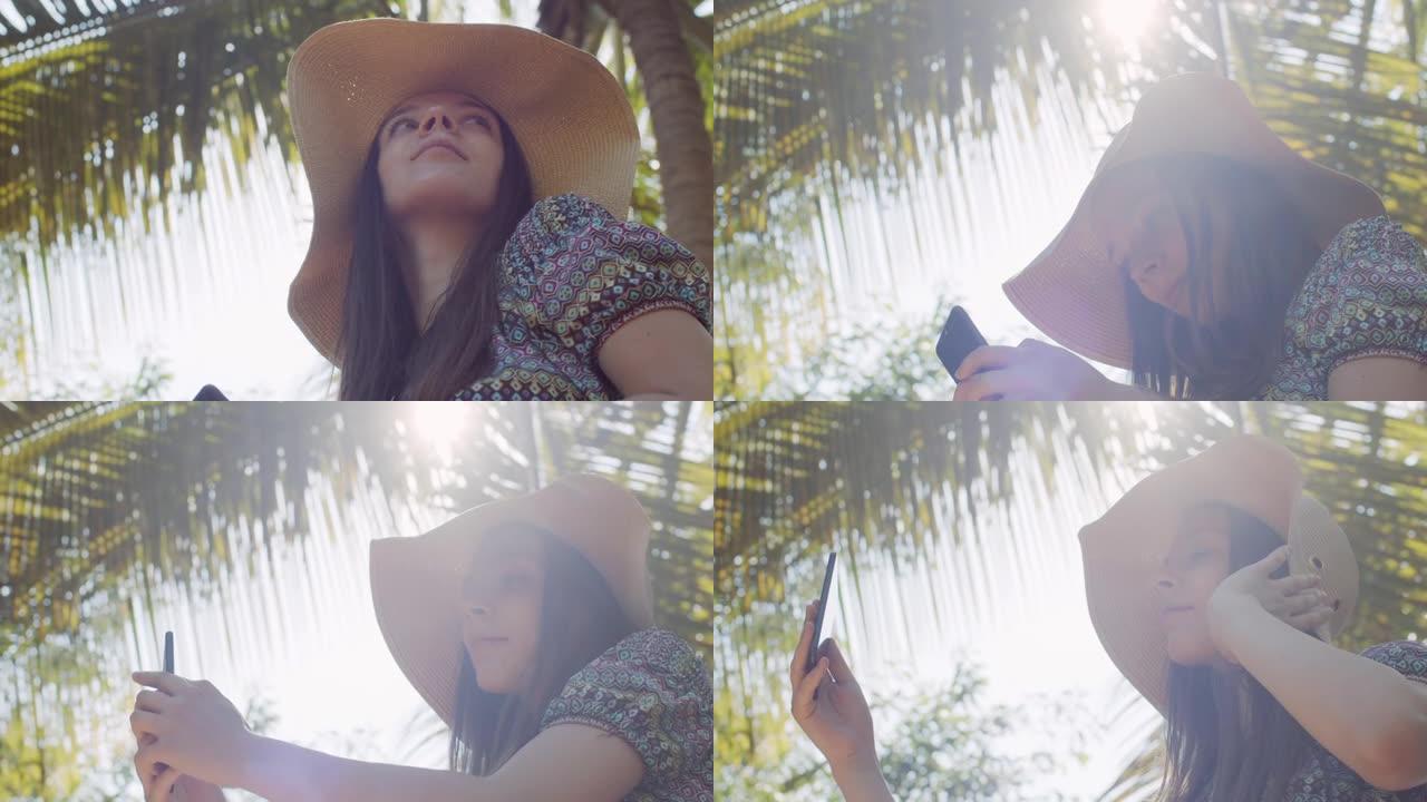 低角度肖像女孩使用手机在热带自然背景。户外肖像优雅的女人在帽子梦想户外手持智能手机镜头光晕。休闲夏日