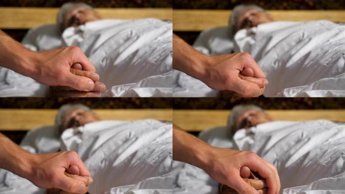 在医疗诊所中，年轻男子握住并安慰生病的成熟女人的手。孙子轻轻地抚摸着躺在病床上的年迈祖母的皱纹手臂。