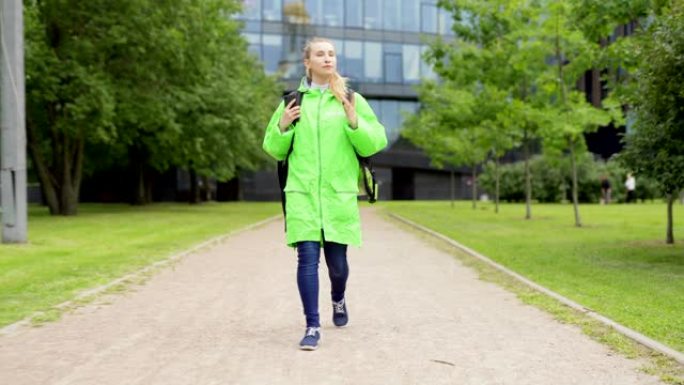 穿着绿色制服和保温袋走在街上的十几岁女性食品快递员的全长前视镜头，在智能手机上使用全球定位系统应用程