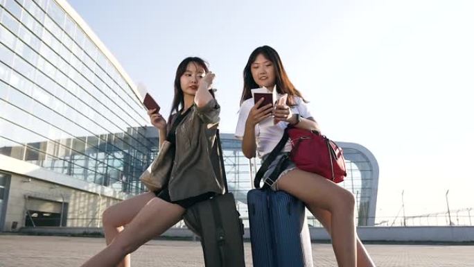 美丽的欢快的25岁亚洲女性最好的朋友坐在行李箱上，手里拿着文件在机场大楼附近飞行