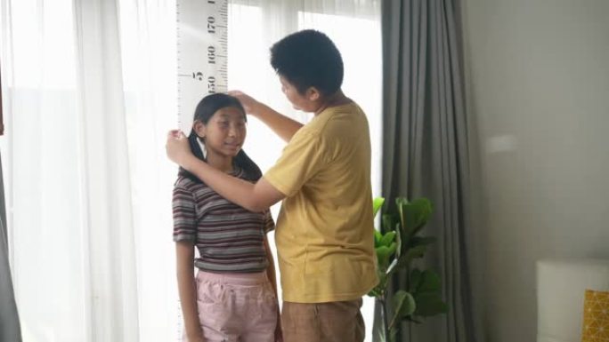快乐的亚洲兄弟在家对着窗户测量他姐姐的身高，生活方式理念。