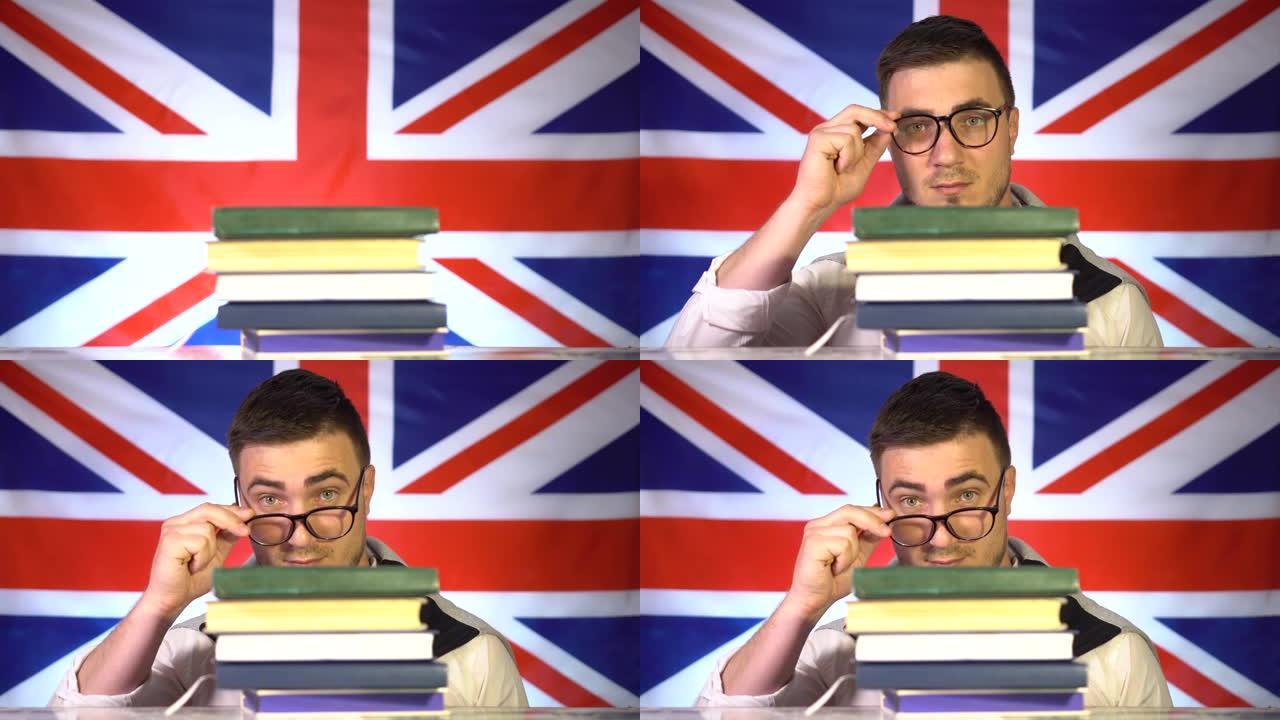 戴着眼镜的可爱的学生家伙从一堆书里偷看出来，微笑着，在英国国旗的背景下，他的眼镜从脸上略微放下。英国